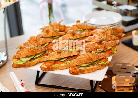 Panini al croissant con salmone affumicato, cetriolo e lattuga disposti su un vassoio bianco a un buffet di catering. Foto Stock