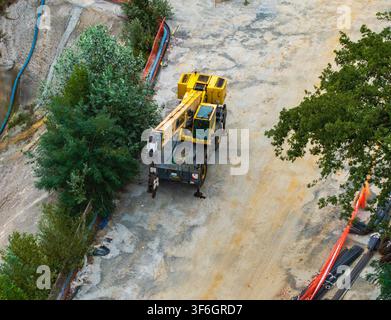 Vista aerea di una gru mobile gialla parcheggiata in un cantiere edile con recinzione di sicurezza arancione, materiali da costruzione e alberi circostanti a Serrav Foto Stock