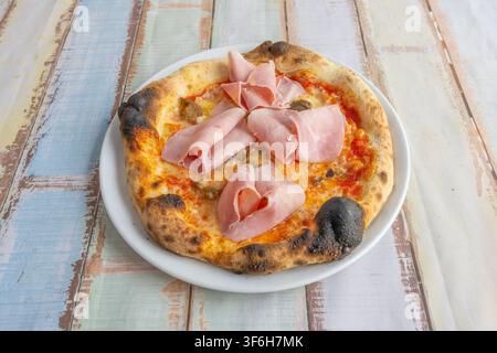 Pizza artigianale bruciacchiata con funghi, formaggio fuso e fette di prosciutto cotto arrotolate, presentata su tavolo di legno rustico pastello Foto Stock