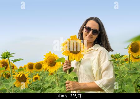 Giovane donna con occhiali da sole che tiene un grande girasole in mezzo a un campo estivo luminoso sotto un cielo blu terso, posa rilassata con luce naturale calda e colori vibranti Foto Stock