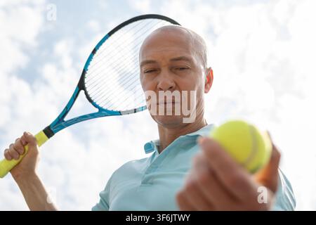 Vista a basso angolo di un giocatore di tennis caucasico che ispeziona la palla da tennis prima del servizio su un campo all'aperto. Concetto di preparazione focalizzata e sport competitivo. Foto Stock