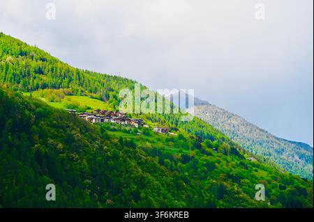 Una fotografia orizzontale di paesaggio di un piccolo insediamento remoto annidato su pendici montuose verdi rigogliose e vibranti, con una vetta rocciosa che si innalza sullo sfondo sotto un cielo nuvoloso. Foto Stock