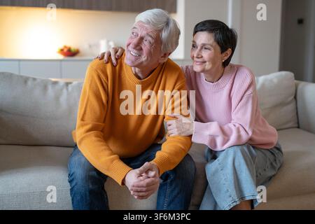 Moglie affettuosa e allegra che abbraccia il marito sorridente mentre la coppia è seduta insieme sul divano a casa. Foto Stock