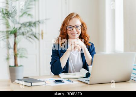 Donna d'affari dai capelli rossi con occhiali sorride davanti a un laptop a una scrivania luminosa in ufficio Foto Stock