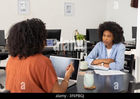 Diverse colleghe donne che esaminano schizzi come pari mostrando un laptop a un tavolo rotondo per il feedback. Collaborazione, ufficio, colleghe, computer, quaderno, diagrammi, discussione Foto Stock