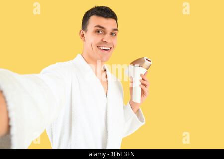 Giovane uomo con fotoepilatore che scatta un selfie su sfondo giallo Foto Stock