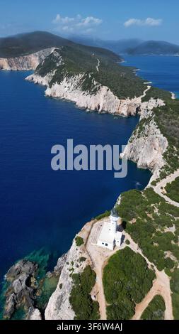 Stupenda ripresa verticale con drone di un faro bianco solitario su una scogliera costiera rocciosa. Bella acqua del mare di un blu profondo, colline verdi, strada sterrata tortuosa, e Foto Stock
