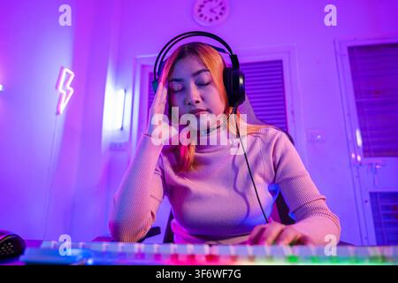 Game over. Gamer asiatico arrabbiato con cuffie da gioco che gioca a un videogioco su PC con luce viola di notte a casa, donna stanca e triste Foto Stock