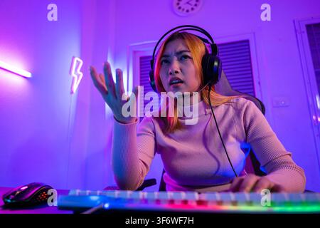Game over. Gamer asiatico arrabbiato con cuffie da gioco che gioca a un videogioco su PC con luce viola di notte a casa, donna stanca e triste Foto Stock