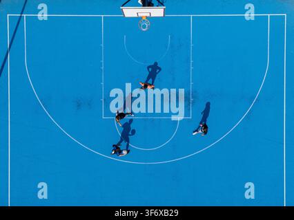 Vista aerea dall'alto di più giocatori in competizione su un campo da basket blu brillante all'aperto con lunghe ombre, concetto di gioco di basket da strada urbano. Foto Stock