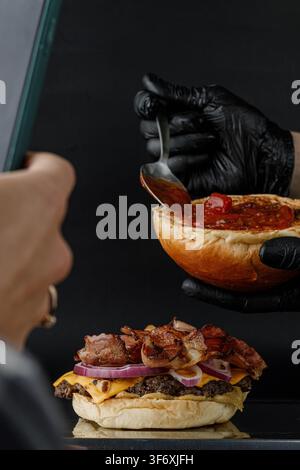 Una persona scatta una foto di uno chef che prepara un burger al bacon Foto Stock