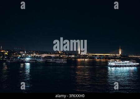 Istanbul, Turkiye - [01.07.2026: Ponte metropolitana del Corno d'Oro illuminata di notte con barche turistiche ormeggiate lungo il lungomare Foto Stock