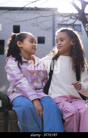Ragazze afroamericane sedute su un basso muretto di cemento, braccio appoggiato sulla spalla, zaini visibili. Controluce, luce di bordo, cortile, urbano, esterno, fri Foto Stock