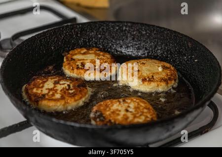 Syrniki dorati e croccanti che cuociono nell'olio bollente dentro una padella di metallo rustica, catturando la texture autentica dei pancake tradizionali di formaggio fresco Foto Stock