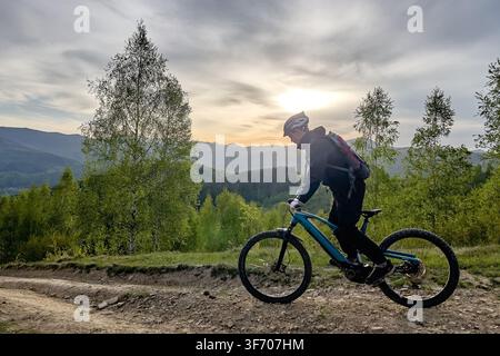 Vista laterale di un mountain biker che pedala su una bici elettrica su un sentiero sterrato attraverso colline boschive verdi al tramonto, paesaggio scenografico e concetto di avventura all'aperto Foto Stock
