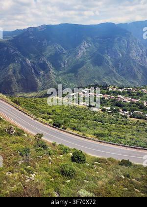 Strada tortuosa si snoda attraverso una valle verdeggiante. Le montagne si ergono sullo sfondo con nuvole morbide. Piccola città incastonata tra alberi e arbusti Foto Stock