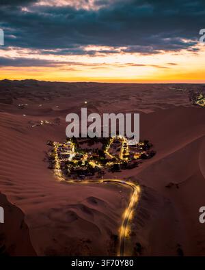 Vista aerea dell'oasi di Huacachina illuminata di notte, circondata da vaste dune di sabbia sotto un drammatico cielo al tramonto a Huacachina, Ica, Perù. Foto Stock