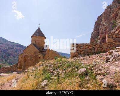 Antico monastero di Noravank in pietra arroccato contro scogliere frastagliate sotto un cielo azzurro terso. Rovine illuminate dal sole si fondono con erba secca e terreno roccioso Foto Stock