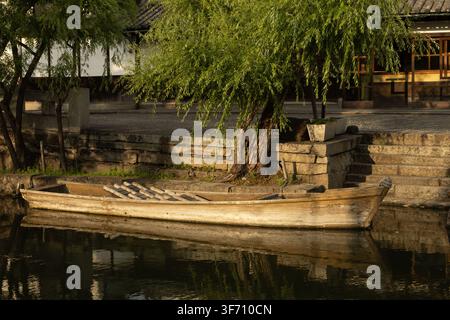 Una barca tradizionale a fondo piatto in legno ormeggiata lungo le rive in pietra fiancheggiate da salici del canale storico di Bikan a Kurashiki, prefettura di Okayama Foto Stock