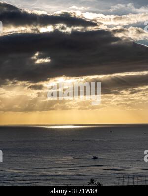 Raggi dorati del crepuscolo si fanno strada attraverso le nuvole di tempesta su un oceano calmo al tramonto, con barche lontane sull'acqua. Foto Stock