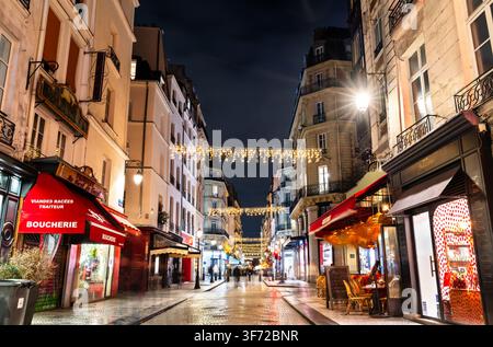 Parigi, Francia – 24 dicembre 2025: Vista stradale di Rue Montorgueil con decorazioni luminose natalizie, vetrine dei negozi e architettura storica illuminata Foto Stock