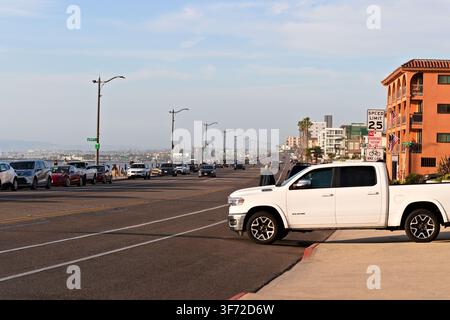 Un pick-up bianco si ferma all'angolo di una strada cittadina. I binari del tram attraversano la strada. Auto parcheggiate costeggiano il marciapiede. Un edificio con tegole rosse si innalza in lontananza. Foto Stock