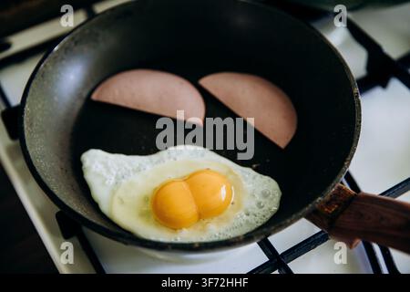 Primo piano di un uovo fritto con tuorlo liquido che viene girato con una spatola in una padella accanto a fette di prosciutto, cotto su un fornello a gas Foto Stock