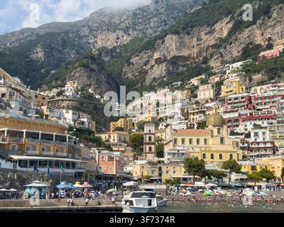 Edifici colorati sulla scogliera e la Chiesa di Santa Maria Assunta si ergono sopra la spiaggia affollata e il porto di Positano sulla Costiera Amalfitana italiana. Foto Stock