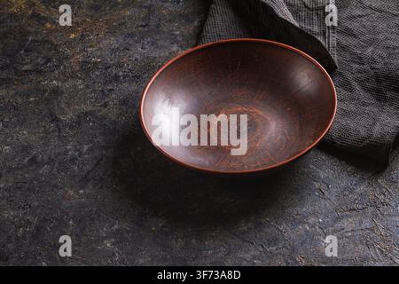 Vista ad angolo alto di una ciotola di terracotta rossa-marrone lavorata a mano vuota e di un tovagliolo in tessuto a maglia waffle grigio scuro su una base rustica in ardesia scura Foto Stock