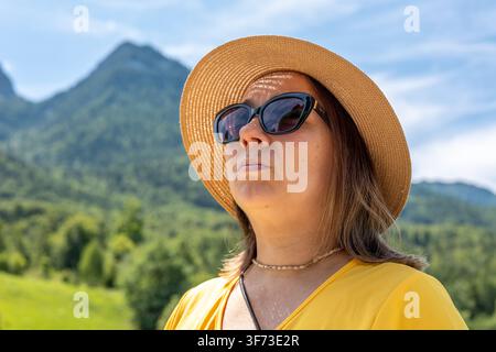 Primissimo piano di una donna di 40 anni che indossa un cappello di paglia e occhiali da sole scuri contro montagne verdi Foto Stock