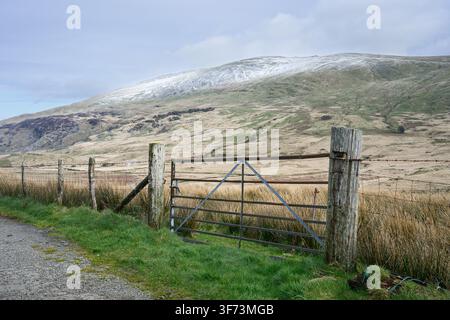 Cancello metallico di fattoria su pali di legno davanti a una montagna innevata e terreni agricoli selvaggi nel nord del Galles Foto Stock