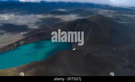 Fotografia aerea di un lago di cratere geotermico con acqua turchese brillante, pendici vulcaniche scure e campi di lava circostanti in. Foto Stock