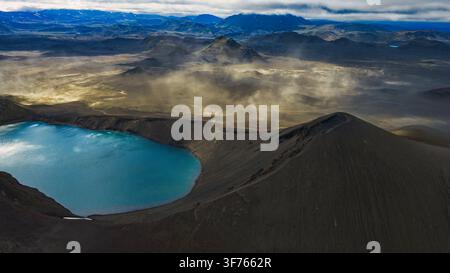 Fotografia aerea di un cratere geotermico con acqua di lago turchese brillante racchiusa da formazioni rocciose vulcaniche scure a Rangárþing. Foto Stock