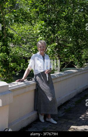 Donna anziana sorridente con capelli corti che indossa occhiali da sole alla moda e si trova vicino a una balaustra in pietra in una giornata estiva soleggiata all'aperto. Foto Stock