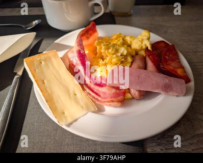 Deliziosa porzione di colazione sostanziosa con strisce di pancetta fritta, uova strapazzate morbide, formaggio a fette, prosciutto saporito, salsicce e caffè sul tavolo. Foto Stock