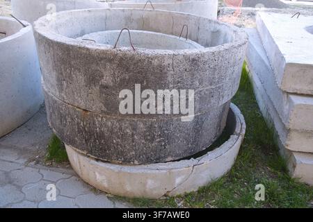 Un grande anello di pozzo nero circolare in cemento è mostrato in un ambiente esterno con erba e una superficie lastricata nelle vicinanze. La struttura sembra consumata e ha Foto Stock