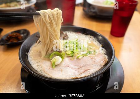 Una ciotola fumante di autentico ramen tonkatsu giapponese con ricco brodo di maiale cremoso, fette di carne tenere, cipollotti e spaghetti sottili sollevati da bacchette. Foto Stock