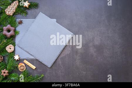 Composizione natalizia con una carta bianca vuota circondata da rami di sempreverdi, pigne, stelle di legno e decorazioni festive su una texture grigio scuro Foto Stock