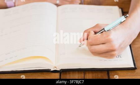 Prima di una mano di donna che scrive in un planner aperto su un tavolo di legno. Ottimo per lifestyle, pianificazione, organizzazione, agenda, journaling e casa Foto Stock