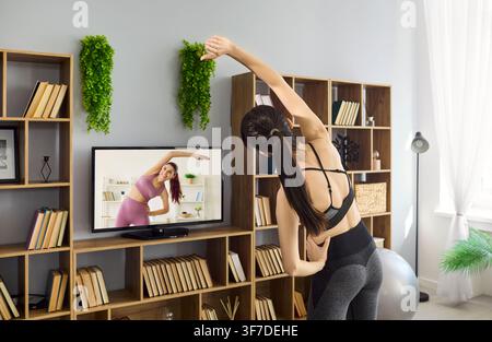 Giovane donna che si allunga a casa con una lezione di fitness online in TV in una stanza moderna e accogliente Foto Stock