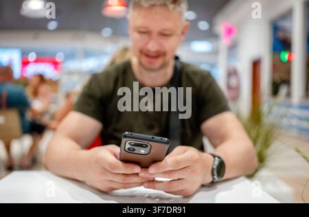 Uomo di mezza età seduto a un tavolo che controlla lo schermo del suo smartphone all'interno di un centro commerciale luminoso Foto Stock