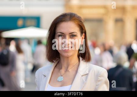 Una donna sorridente in un blazer beige si trova all'aperto con una folla cittadina sfocata sullo sfondo. Foto Stock