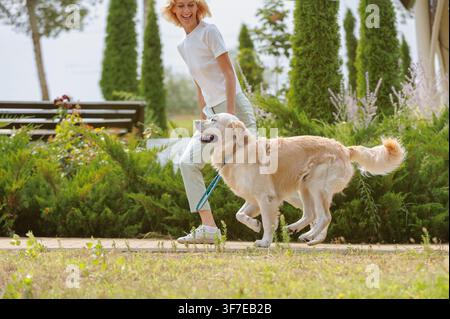 Donna felice che gioca, salta, si diverte con un cane Golden Retriever al guinzaglio in un parco urbano, stile di vita all'aperto gioioso, compagnia attiva, estate verde Foto Stock