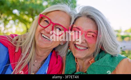 Amiche anziane sorridono e ridono, indossano occhiali colorati all'aperto Foto Stock