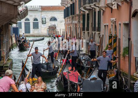 ITALIA, VENEZIA, 11 luglio 2025: Diversi gondolieri in camicie a righe navigano gondole nere con passeggeri lungo un canale stretto fiancheggiato da edifici storici Foto Stock