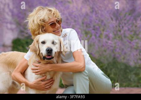 Donna felice con occhiali da sole che abbraccia un Golden Retriever, sorride, godendosi uno stile di vita all'aperto, lavanda, fiori viola, cane che guarda l'obiettivo, spazio libero Foto Stock