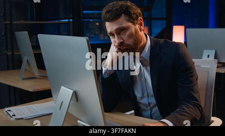 Uomo d'affari caucasico annoiato e demotivato che lavora con noiosi compiti al computer in ufficio di notte, la sera, triste, frustrato, stressato, oberato di lavoro, di mezza età Foto Stock