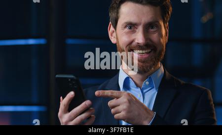 Uomo d'affari caucasico di mezza età sorridente che indica il telefono cellulare in un ufficio notturno uomo d'affari maturo capo uomo dirigente che tiene uno smartphone Foto Stock