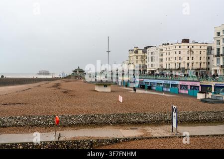 I turisti godono di una passeggiata tranquilla lungo Brighton Beach, nell'East Sussex, passando davanti alla torre i360 e alle rovine del Palace Pier in una giornata calma e nuvolosa. Foto Stock