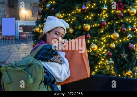 Donna felice che si gode gli acquisti natalizi in città con un albero festivo Foto Stock